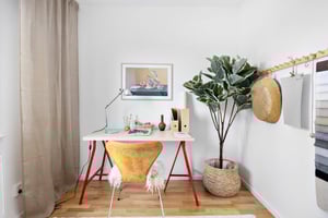 Arbetsyta med ett vitt skrivbord på röda ben, en gul stol med fårskinn, en bordslampa, dekorationsföremål och en stor grön växt i en flätad kruka