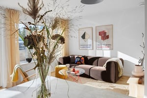 Vardagsrum med en brun soffa, soffbord och dekorativa affischer på väggen. En vas med torkade växter står på ett matbord i förgrunden