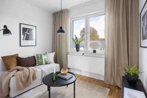 Vardagsrum med ljusa väggar, en vit soffa med kuddar och filt, ett runt soffbord, gröna växter och stora fönster med beige gardiner