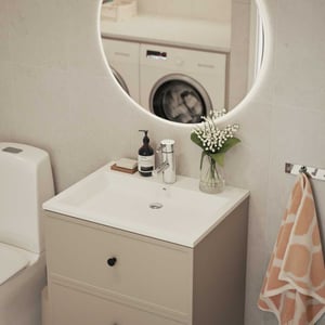 Modernt badrum med vit toalett, beige handfatsskåp, rund spegel med belysning, blomvas och tvättområde synligt i bakgrunden