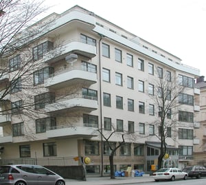 Vit bostadsbyggnad med flera våningar och balkonger, träd i förgrunden och parkerade bilar vid trottoaren