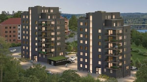 Två moderna lägenhetsbyggnader med mörk fasad och flera våningar, omgivna av grönska och parkerade bilar, med en sjö och skog i bakgrunden