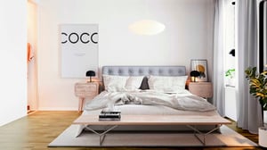 Sovrum med en grå stoppad sängram, vita sängkläder och nattduksbord på båda sidor. Väggdekoration med texten "COCO", golvlampa och grön växt vid fönstret