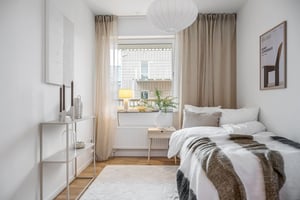 Sovrum med vit inredning, stor fönster med beige gardiner, säng med vit och grå bäddning, vit taklampa och dekorativ hylla