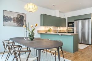 Modernt kök och matplats med gröna skåp, köksö, svart matbord, trästolar och tulpaner i en vas. Väggkonst och hängande lampa syns ovanför bordet