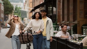 Tre personer promenerar på en trottoar bredvid ett kafé, där en person sitter vid ett bord; en av dem leder en cykel