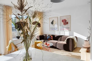 Vardagsrum med en brun soffa, soffbord och dekorativa affischer på väggen. En vas med torkade växter står på ett matbord i förgrunden
