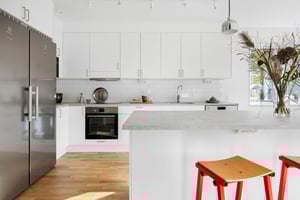 Modernt kök med vita skåp, rostfria vitvaror, ljus bänkskiva och röda barstolar vid köksön