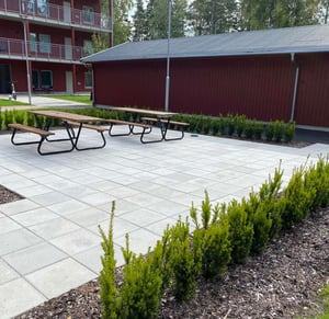 Uteplats med bänkbord, stenlagd gård och röda bostadshus med grönska runtomkring