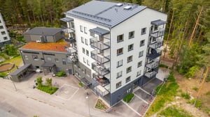 Flygfoto över ett bostadshus med vita fasader, balkonger och skogsomgivning