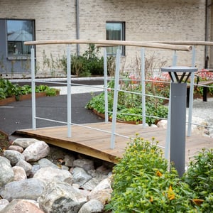 Trädgårdsbro med träbana, vita räcken och omgivande planteringar vid ett bostadshus