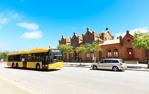 Gul stadsbuss och silverfärgad bil framför en röd tegelbyggnad under klarblå himmel