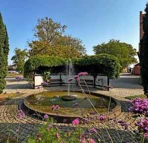 Brunn med fontän i en grönskande innergård omgiven av blommor och kullersten