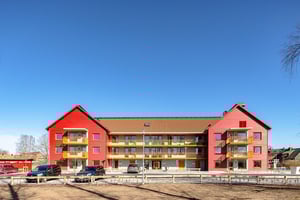 Rött flerbostadshus med balkonger och parkerade bilar framför under klarblå himmel