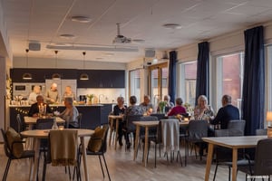 Människor som sitter och står vid bord i en gemensam matsal med kök, lampor och stora fönster