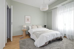 Sovrum med bäddad dubbelsäng, ljusa väggar, gardiner och en liten sidobordslampa