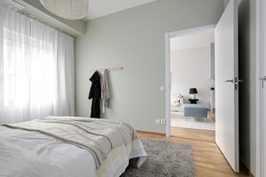 Sovrum med säng, ljusgröna väggar och naturligt ljus från stort fönster. Genom den öppna dörren syns ett ljust vardagsrum