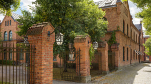 Rött tegelhus med historisk arkitektur bakom en smidesgrind och lummiga träd