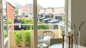 Ljust vardagsrum med öppen dörr ut mot en balkong och utsikt över bostadshus och gröna innergårdar