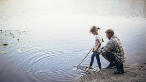 Man och barn vid en sjö, där barnet står i vattnet med en pinne vid strandkanten