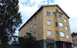 Flerbostadshus i beige fasad med butikslokaler i bottenplan och träd vid sidan