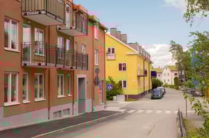 Flerbostadshus i orange och gult längs en lugn gata med balkonger, parkerade bilar och gröna träd
