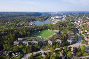 Flygvy över ett bostadsområde med grönområden, fotbollsplan och sjö i bakgrunden