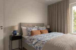 Sovrum med bäddad dubbelsäng, nattduksbord med lampa och fönster med beige gardiner