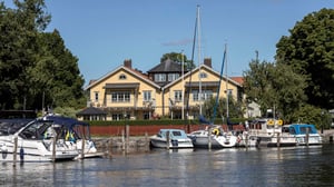 Gul sekelskiftesvilla i Trosa vid vattnet med förtöjda båtar i förgrunden och grönskande träd runtom