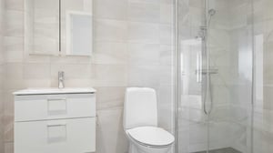 Inflyttningsklart badrum med vita kakelväggar, kommod, toalett och dusch med glasvägg