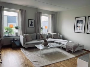 Ljust vardagsrum med hörnsoffa, soffbord i glas och väggdekorationer. Rummet har stora fönster, gardiner och en ljus matta på parkettgolv