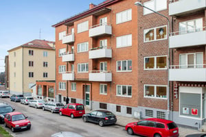 Gatubild med flera flerbostadshus i tegel, parkerade bilar längs trottoaren och klar blå himmel i bakgrunden