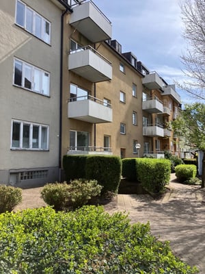 Gul flerfamiljsbyggnad med vita balkonger omgiven av grönska och en gångväg under en solig himmel