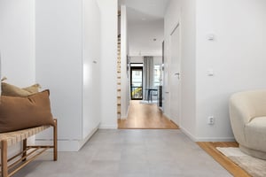 Hall med ljusa vita väggar, klinkergolv, en träbänk med kuddar till vänster och en beige fåtölj till höger. Utsikt mot en balkong genom glasdörrar i bakgrunden