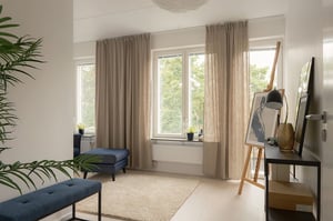 Inflyttningsklar lägenhet med ljust vardagsrumsmiljö, stora fönster, beige gardiner och blå sittbänkar