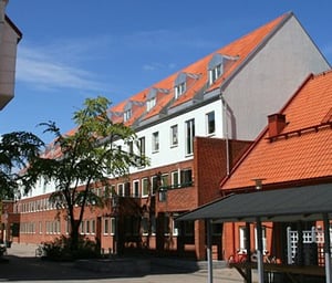 Byggnad med tegeltak och vita fasader under en klarblå himmel, omgiven av träd och en gård