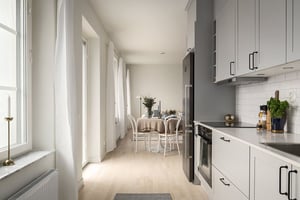 Modernt kök med vita skåp och rostfria vitvaror, anslutet till en matplats med ett dukat bord och stolar. Ljusa trägolv och stora fönster som släpper in naturligt ljus