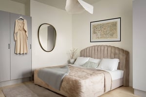 Sovrum med en beige säng, kudde och filt, en spegel på väggen, en inramad karta, och en beige klänning som hänger på en grå garderob
