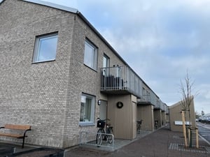 Låga byggnader med fasader av grått tegel, balkonger och cyklar parkerade framför. Vintertid med molnig himmel och torra markytor