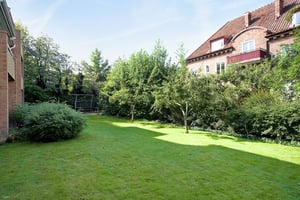 Grön gräsmatta omgiven av buskar och träd, med bostadshus i bakgrunden