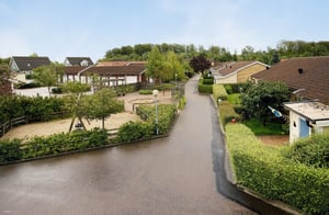 Bostadsområde med villa och radhus, omgiven av grönska och små trädgårdar, med en asfalterad väg i mitten under en klar himmel