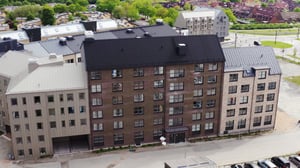 Flerfamiljshus med fasader i olika nyanser av brunt och grått, omgiven av grönska och parkeringsplatser