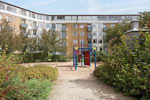 Lekplats med sand och lekutrustning omgiven av buskar, träd och ett flerbostadshus i bakgrunden