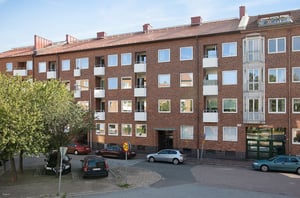 Byggnad med fasad av röd tegel, flera våningsplan och vita balkonger, omgiven av parkerade bilar och grönska