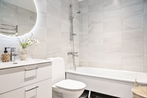 Modernt badrum med vita kakelväggar, rund spegel med belysning, toalett, badkar och handfat med dekorativ vas och tvålpump