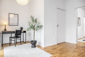 Ljus inredning med ett skrivbord, stol och bordslampa bredvid en växt i kruka på ett vitt matta. Väggarna är vita, och dörrar leder till olika rum