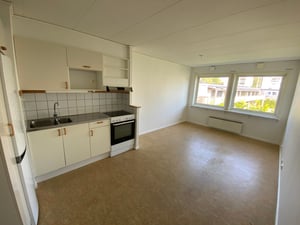 Kök och vardagsrum i en lägenhet med vita väggar, ljusbrunt golv, och fönster som vetter mot en innergård. Köksdelen har vita skåp, vit kyl och spis med svart häll samt kakel som stänkskydd