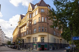 Gulbrunt flerfamiljshus i tegel med spetsiga tak, stora fönster och butiker på bottenvåningen, beläget vid en gatukorsning med omgivande grönska och parkeringsskylt