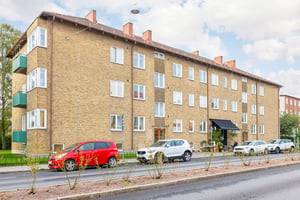 Brun tegelbyggnad med flera våningar, fönster och balkonger, bilar parkerade framför på en asfalterad väg