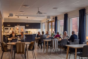 Människor som sitter och står vid bord i en gemensam matsal med kök, lampor och stora fönster